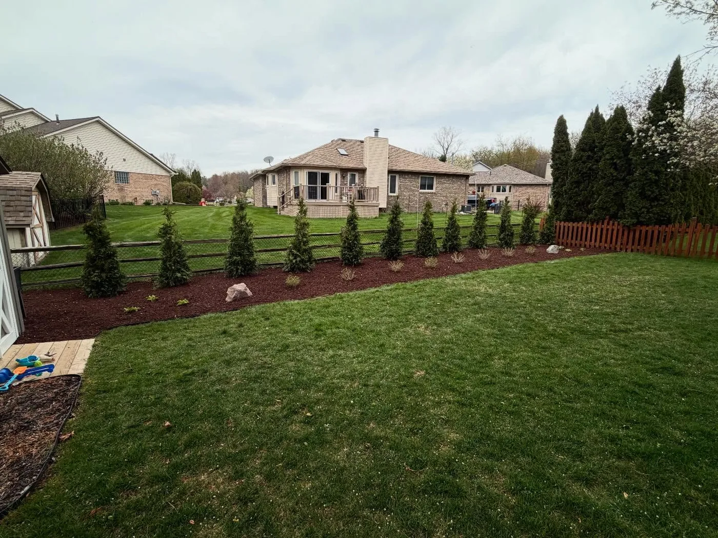 Finished Big Dawgs privacy-row installation with evergreens, flowering shrubs, boulders, and a curved mulch bed